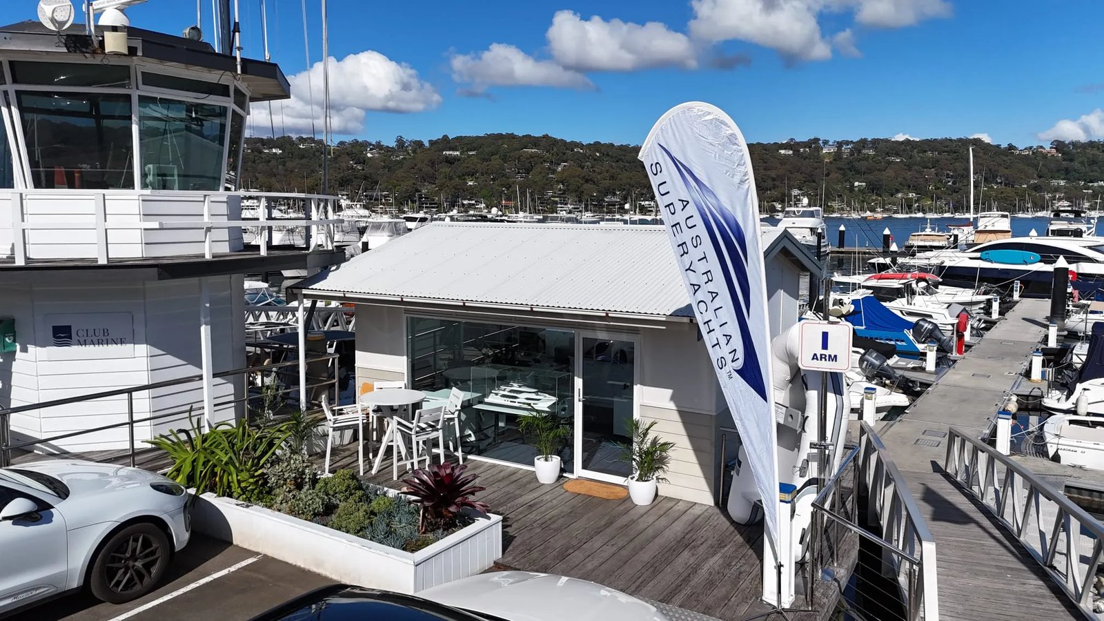 The Australian Superyachts office with its flag on the deck at the Royal Motor Yacht Club, Newport, with boats on their berths and the wooded shore of Pittwater behind
