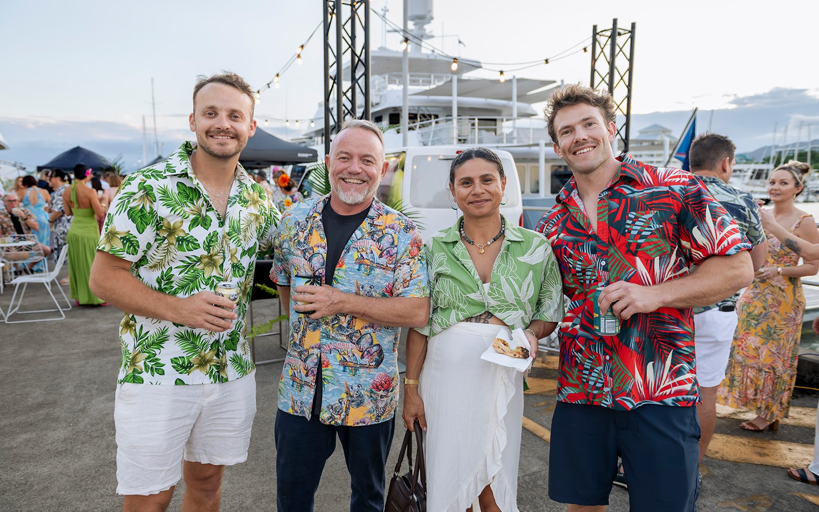Four smiling guests in tropical shirts and summer clothes on Sailfish Quay at dusk, with a superyacht, festoon lights and more guests behind them